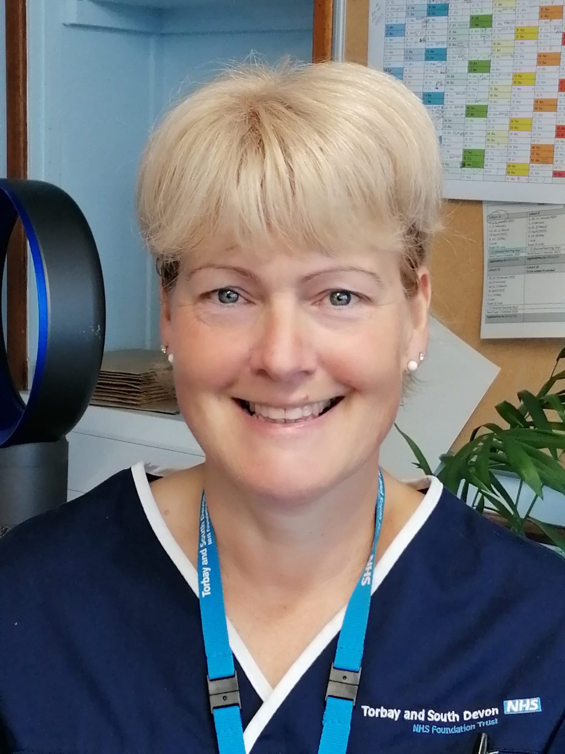 Becky, a white woman, has short blonde hair and is wearing pearl earrings. She is smiling and wearing an NHS lanyard and dark blue nurses uniform.